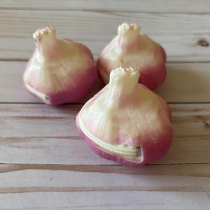 May include: Three novelty onion-shaped zippered pouches. The pouches are white and pink, with a zipper closure. The pouches are arranged on a light-colored wooden surface.
