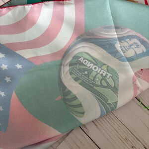 May include: A folded fabric featuring the flags of the United States, Canada, and a green and white design. The American flag has red and white stripes with blue stars. The Canadian flag is red and white. The green and white design has text.