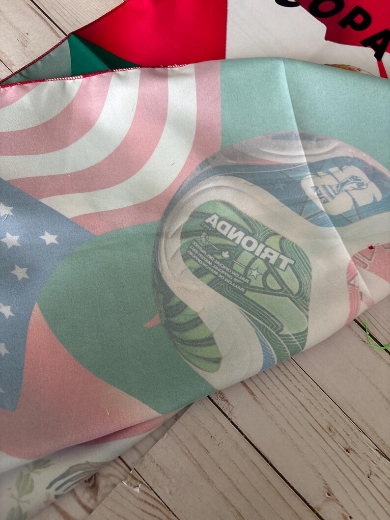 May include: A folded fabric featuring the flags of the United States, Canada, and a green and white design. The American flag has red and white stripes with blue stars. The Canadian flag is red and white. The green and white design has text.