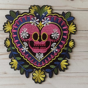 May include: Embroidered patch featuring a pink sugar skull design within a heart shape. The patch is adorned with colorful floral and leaf accents in yellow, blue, and white, set against a black background.