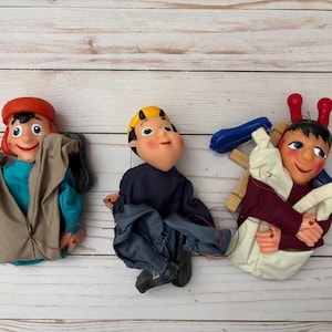 May include: Three hand puppets with different facial expressions and clothing. The puppet on the left is wearing a brown and blue outfit with a red hat. The puppet in the middle is wearing a blue and grey outfit with a yellow hat. The puppet on the right is wearing a white and red outfit with a blue and red hat.