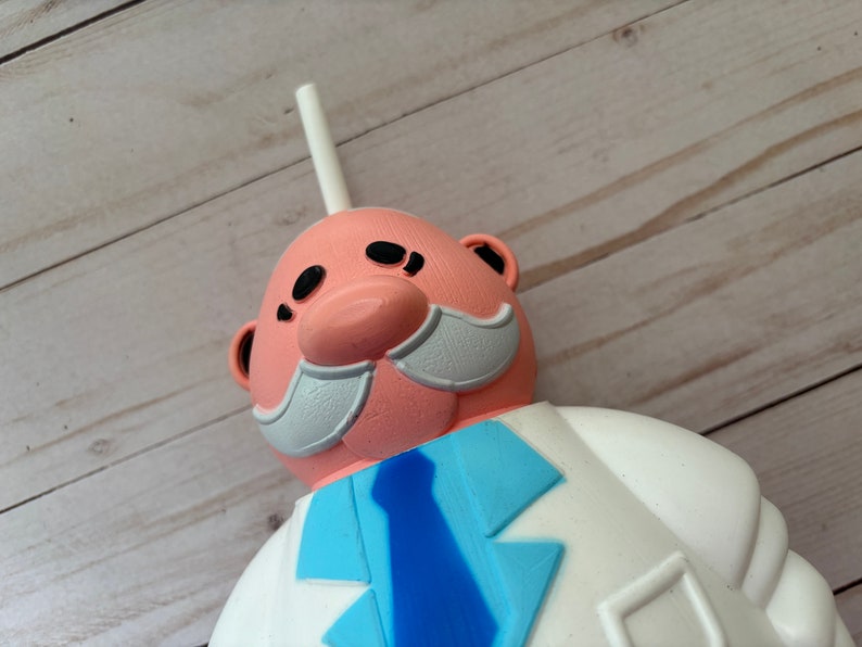Peut inclure: Un gobelet en plastique rose et blanc en forme de personnage de dessin anim&eacute; avec une paille blanche. Le personnage a une moustache blanche, une cravate bleue et une blouse de laboratoire blanche.