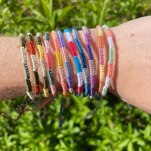 May include: A collection of colorful friendship bracelets. Each bracelet is made of woven threads in various colors, including green, red, blue, yellow, and pink. The bracelets are worn on a wrist against a background of green foliage.