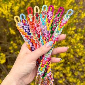 Może przedstawiać: Kolekcja kolorowych bransoletek przyjaźni ze skomplikowanymi wzorami tkanymi. Każda bransoletka ma inny schemat kolorów, w tym różowy, żółty, niebieski i zielony. Bransoletki są trzymane w dłoni na tle żółtych kwiatów.