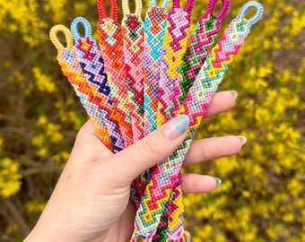Puede incluir: Una colección de siete pulseras de la amistad tejidas a mano con intrincados patrones de chevrón y diamante en vibrantes combinaciones de hilos multicolores que incluyen rosa, amarillo, azul, verde, naranja y rojo, cada una con un lazo.