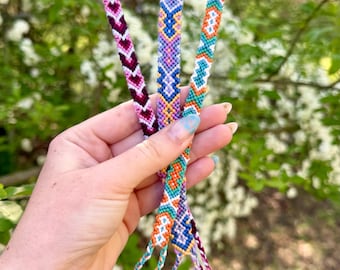 Puede incluir: Tres pulseras de la amistad tejidas con intrincados patrones geométricos. Una pulsera muestra chevrones en color burdeos y blanco, otra rombos en lavanda y azul, y la tercera presenta motivos en naranja vibrante, turquesa y blanco.