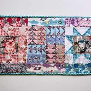 May include: A patchwork quilt runner with a teal border. The quilt features a variety of floral and geometric patterns in pink, blue, and white. The center of the quilt features a pig and a cow.