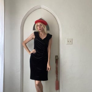 Puede incluir: Un vestido de terciopelo negro con un escote de volantes. El vestido es de largo hasta la rodilla y tiene una silueta ajustada. La modelo lleva una boina roja.