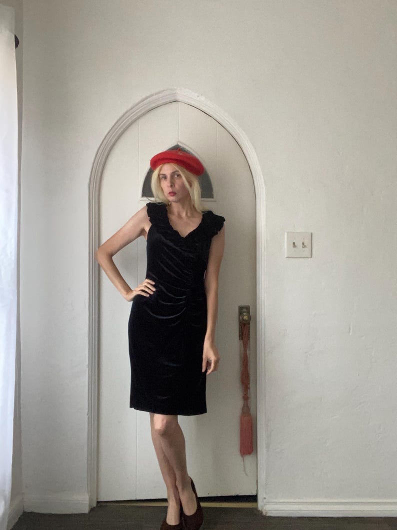 Puede incluir: Un vestido de terciopelo negro con un escote de volantes. El vestido es de largo hasta la rodilla y tiene una silueta ajustada. La modelo lleva una boina roja.