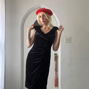 Puede incluir: Una mujer con un vestido de terciopelo negro con un escote de volantes y una boina roja. Est&aacute; de pie frente a una puerta blanca con un borla rosa.