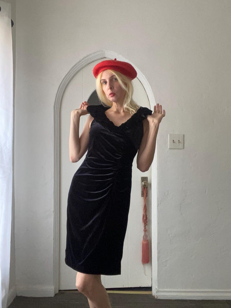 Puede incluir: Una mujer con un vestido de terciopelo negro con un escote de volantes y una boina roja. Est&aacute; de pie frente a una puerta blanca con un borla rosa.
