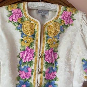 May include: Ivory-colored floral jacket with a decorative floral design in pink, blue, and yellow. The jacket has gold trim and small gold buttons. The label reads "Adrianna Papell".