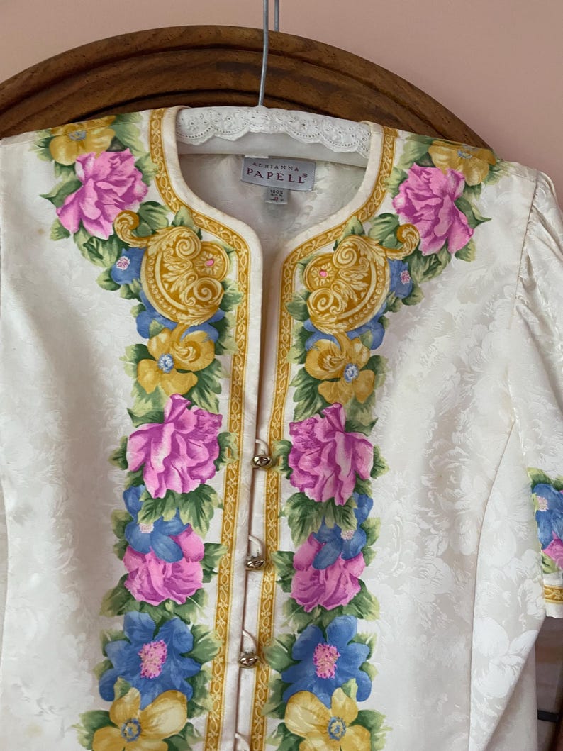 May include: Ivory-colored floral jacket with a decorative floral design in pink, blue, and yellow. The jacket has gold trim and small gold buttons. The label reads "Adrianna Papell".