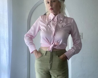 May include: A woman wearing a pink satin western-style shirt with silver star embellishments, tied at the waist, and green plaid pants. She is wearing a maroon beret.