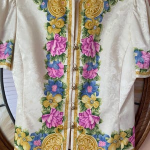 May include: Ivory-colored jacket with a floral design. The jacket features a pattern of pink, blue, and yellow flowers with gold accents. It has short sleeves and decorative gold buttons down the front. The brand name "Adrianna Papell" is visible.
