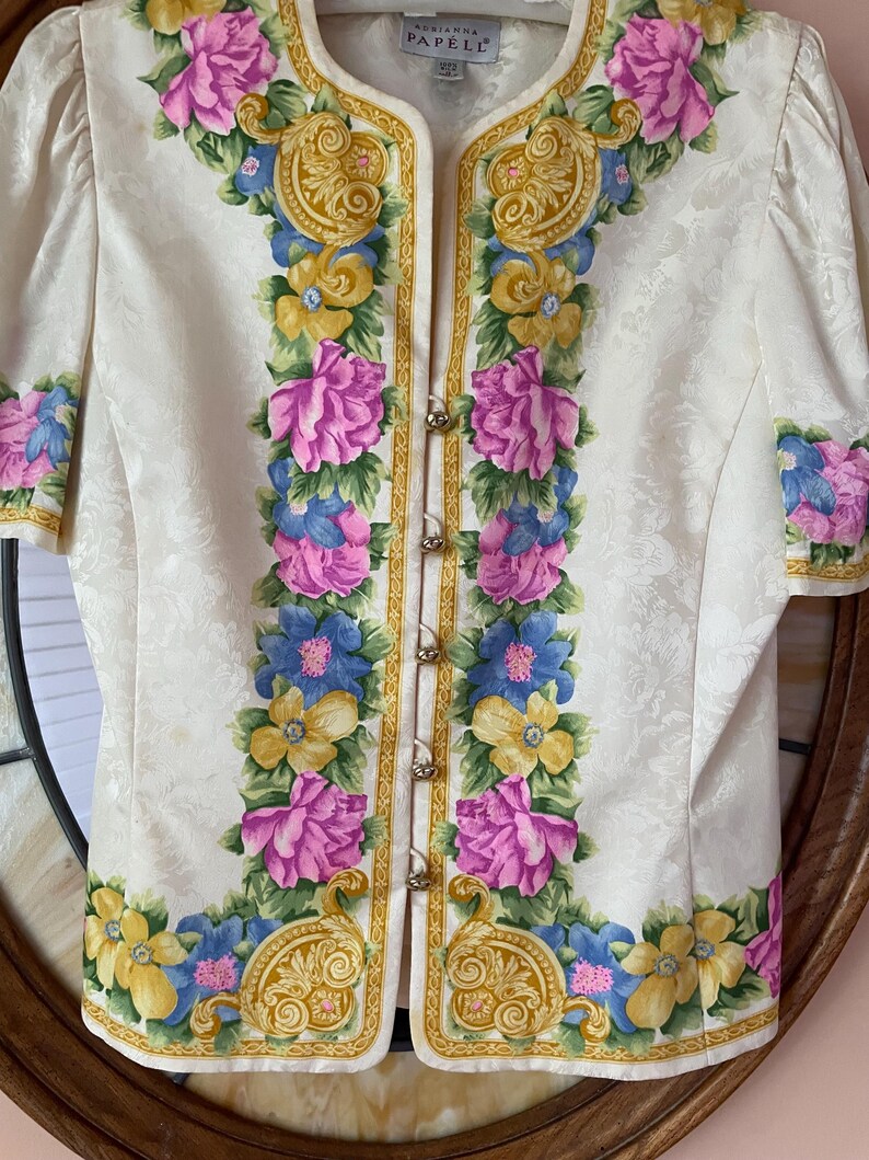 May include: Ivory-colored jacket with a floral design. The jacket features a pattern of pink, blue, and yellow flowers with gold accents. It has short sleeves and decorative gold buttons down the front. The brand name "Adrianna Papell" is visible.
