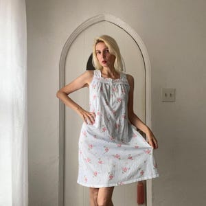 May include: A sleeveless, knee-length nightgown in white with a delicate floral pattern of pink roses and green leaves. The nightgown features a square neckline and a button-up front, offering a vintage aesthetic.