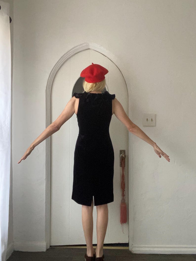 Puede incluir: Una mujer con un vestido de terciopelo negro y una boina roja. El vestido tiene un escote con volantes y una silueta ajustada.