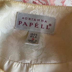May include: Close-up of a cream-colored silk garment label. The label reads "ADRIANNA PAP&Eacute;LL" in a serif font. Below, it states "100% Silk" and "Made in Korea". The garment has a subtle floral pattern.