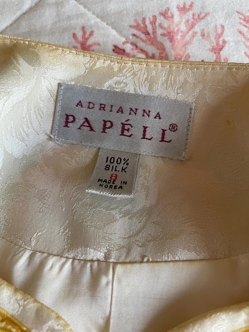 May include: Close-up of a cream-colored silk garment label. The label reads "ADRIANNA PAP&Eacute;LL" in a serif font. Below, it states "100% Silk" and "Made in Korea". The garment has a subtle floral pattern.