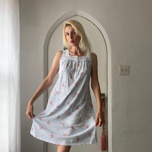 May include: A light blue sleeveless dress with a floral pattern. The dress features a square neckline and a button-down front. Pink roses are scattered across the fabric. The dress is knee-length and has a relaxed fit.