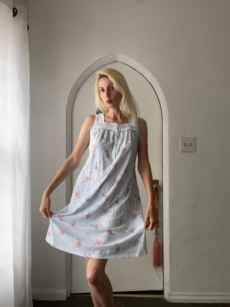 May include: A light blue sleeveless dress with a floral pattern. The dress features a square neckline and a button-down front. Pink roses are scattered across the fabric. The dress is knee-length and has a relaxed fit.