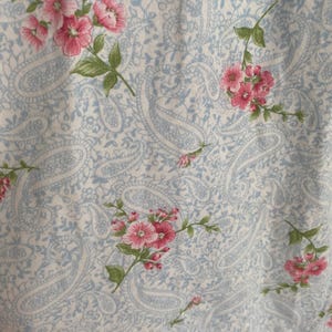 May include: Close-up of a fabric with a light blue paisley pattern and scattered pink floral sprigs. The flowers have multiple petals and green leaves. The fabric appears to be soft and possibly cotton.