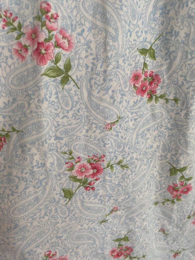 May include: Close-up of a fabric with a light blue paisley pattern and scattered pink floral sprigs. The flowers have multiple petals and green leaves. The fabric appears to be soft and possibly cotton.