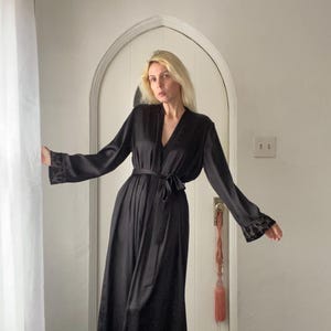 Pode incluir: Uma longa bata de cetim preto com decote em V e mangas compridas. A bata tem um cinto para amarrar e detalhes de renda nas mangas.