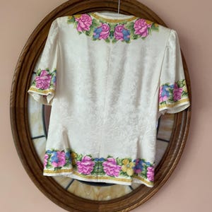 May include: Cream-colored blouse with short sleeves, adorned with a floral pattern. The neckline, sleeve cuffs, and hem are embellished with pink, blue, and yellow floral designs. The blouse is hanging on a metal hanger.
