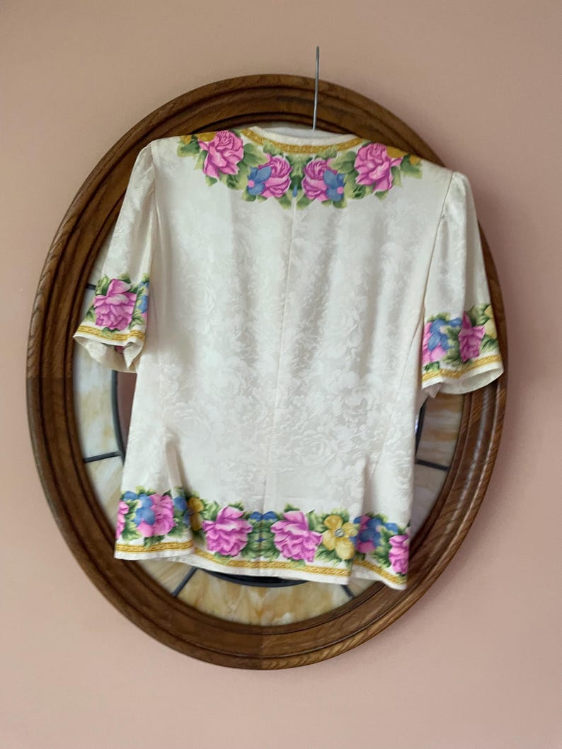 May include: Cream-colored blouse with short sleeves, adorned with a floral pattern. The neckline, sleeve cuffs, and hem are embellished with pink, blue, and yellow floral designs. The blouse is hanging on a metal hanger.