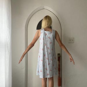 May include: A light blue sundress with a floral pattern of pink roses. The dress has a square neckline and a simple, A-line silhouette. The fabric appears to be lightweight, suitable for warm weather.