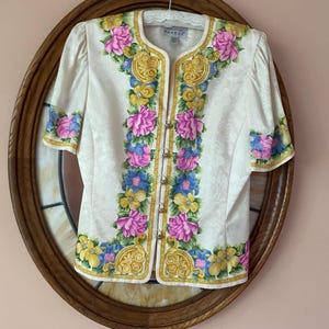 May include: Cream-colored short-sleeved blouse with floral embroidery in pink, blue, and yellow. The blouse has gold-colored trim and small gold-colored buttons. The blouse is displayed on a wooden oval frame.