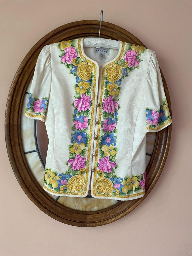 May include: Cream-colored short-sleeved blouse with floral embroidery in pink, blue, and yellow. The blouse has gold-colored trim and small gold-colored buttons. The blouse is displayed on a wooden oval frame.