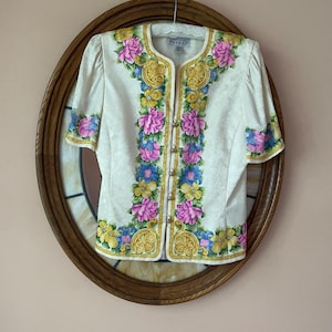 May include: Cream-colored short-sleeved blouse with a floral design in pink, blue, and yellow. The blouse has gold trim and small gold buttons down the front. The blouse is hanging on a hanger against a brown oval mirror.