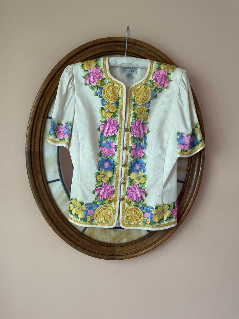 May include: Cream-colored short-sleeved blouse with a floral design in pink, blue, and yellow. The blouse has gold trim and small gold buttons down the front. The blouse is hanging on a hanger against a brown oval mirror.