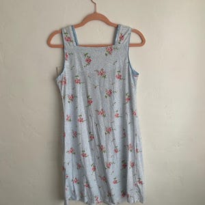 May include: A light blue, sleeveless dress with a square neckline and a floral pattern of pink roses. The dress is hanging on a pink hanger against a white wall. The dress appears to be made of a lightweight fabric.
