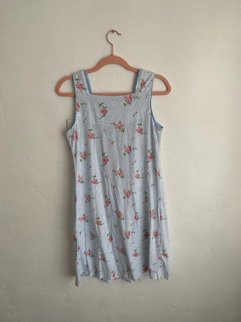 May include: A light blue, sleeveless dress with a square neckline and a floral pattern of pink roses. The dress is hanging on a pink hanger against a white wall. The dress appears to be made of a lightweight fabric.