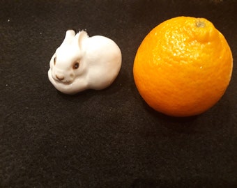 以下が含まれることがあります： 茶色の目をした白いセラミック製のウサギの置物と小さなオレンジ。ウサギは休息のポーズをとっています。オレンジは丸く、皮はざらざらしています。アイテムは暗い表面にあります。
