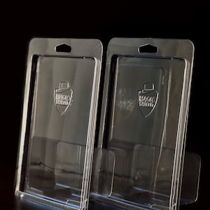 May include: Two clear plastic card holders with a shield logo that reads "MAGIC SHIELD". Each holder has a rectangular shape with a small hole at the top and a stand at the bottom. The holders are on a dark surface.