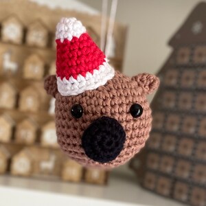 Könnte beinhalten: Ein braunes gehäkeltes Tier, das eine rot-weiße gehäkelte Weihnachtsmütze trägt. Das Tier hat schwarze Augen und eine schwarze Nase.