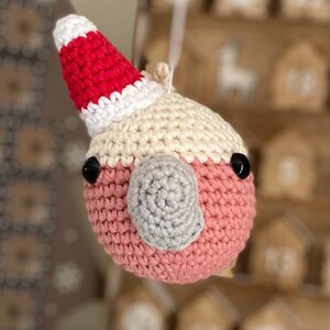 May include: A crocheted pink and white bird wearing a red and white Santa hat. The bird has black eyes and a gray beak.