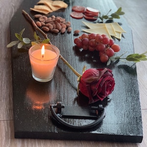 May include: A black serving tray with a variety of snacks, including crackers, cheese, salami, grapes, and a lit candle. A red rose is also on the tray.