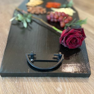May include: A black wooden serving tray shaped like a coffin with a black metal handle. The tray is decorated with grapes, red pepper flakes, nuts, and a single red rose.