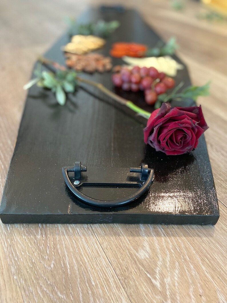 May include: A black wooden serving tray shaped like a coffin with a black metal handle. The tray is decorated with grapes, red pepper flakes, nuts, and a single red rose.