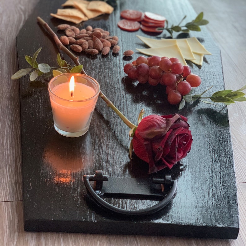 May include: A black serving tray with a variety of snacks, including crackers, cheese, salami, grapes, and a lit candle. A red rose is also on the tray.
