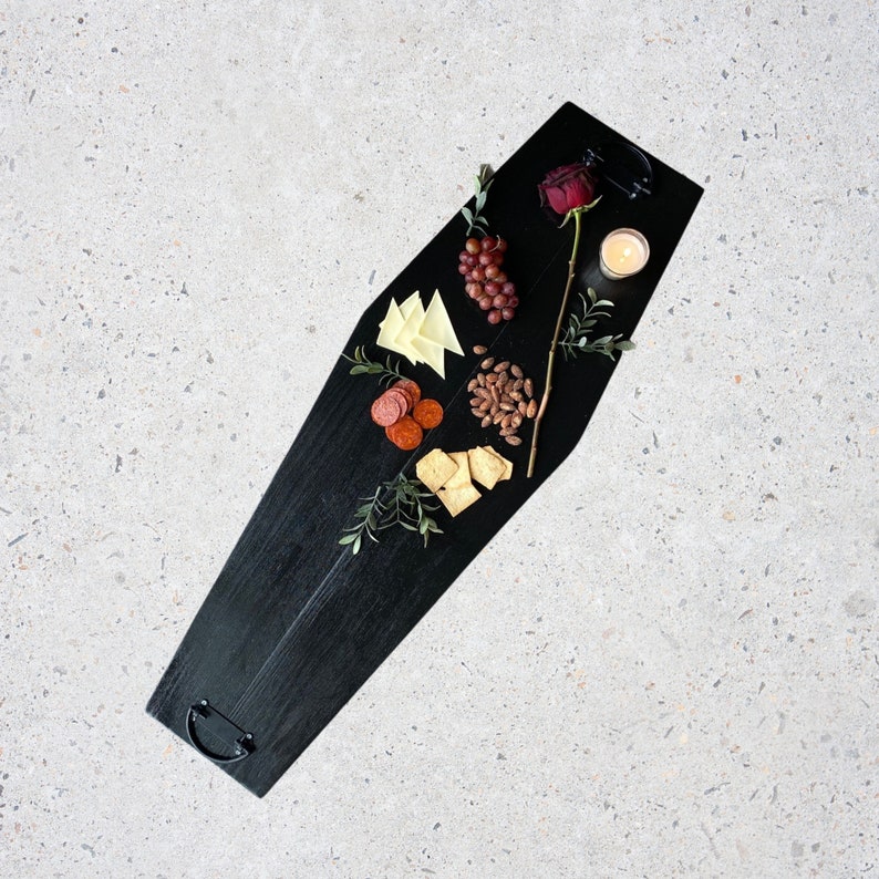 May include: A black coffin-shaped serving tray with a variety of snacks, including cheese, crackers, salami, nuts, grapes, and a single red rose.
