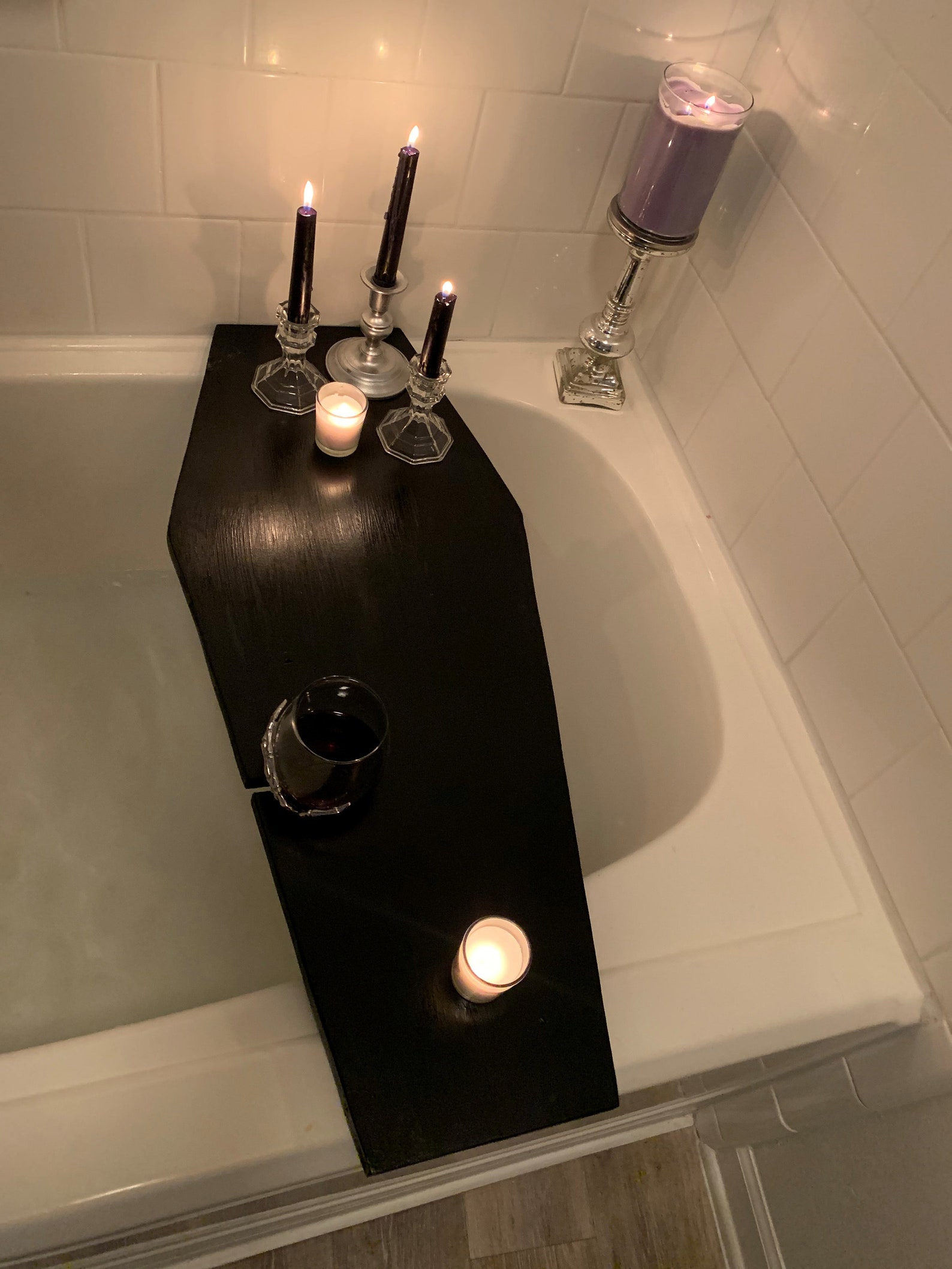 Black coffin-shaped bath tray on white tub, topped with candles and a glass of wine for a gothic soak