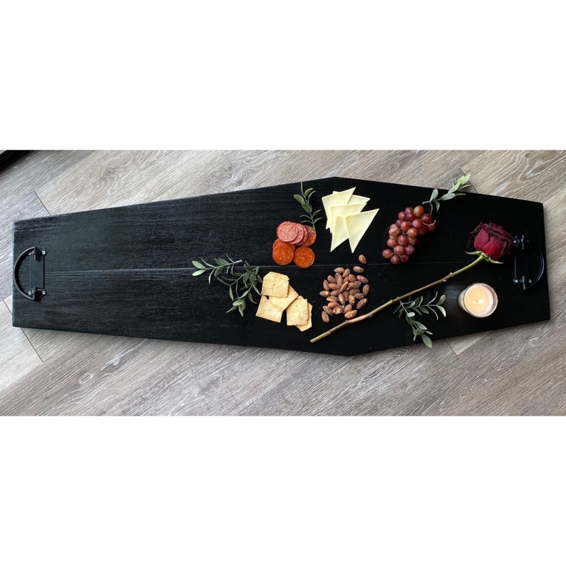 May include: A black wooden serving tray shaped like a coffin with handles on each end. The tray is filled with snacks, including crackers, cheese, pepperoni, almonds, grapes, and a single red rose.