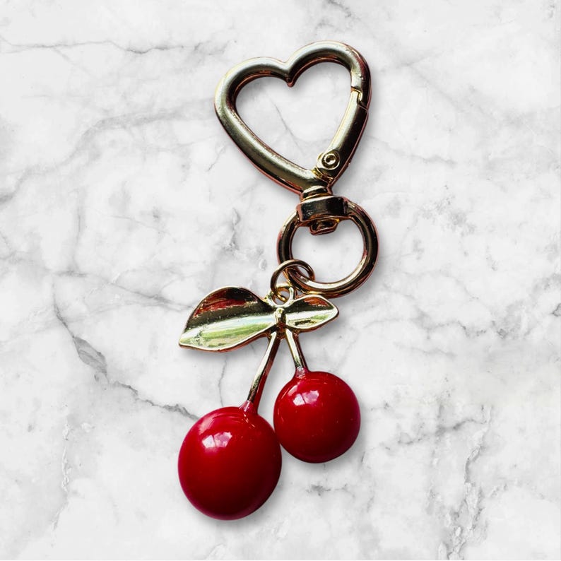 Op de afbeelding: Een goudkleurige sleutelhanger met een hartvormige sluiting en een bedel met twee rode kersen met groene bladeren. De sleutelhanger is ontworpen om aan sleutels of tassen te bevestigen.
